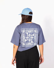 A person shown from the back wearing a light blue baseball cap, a slate blue graphic t-shirt with a large white bandana-style print, and black wide-leg pants, standing against a white studio background.