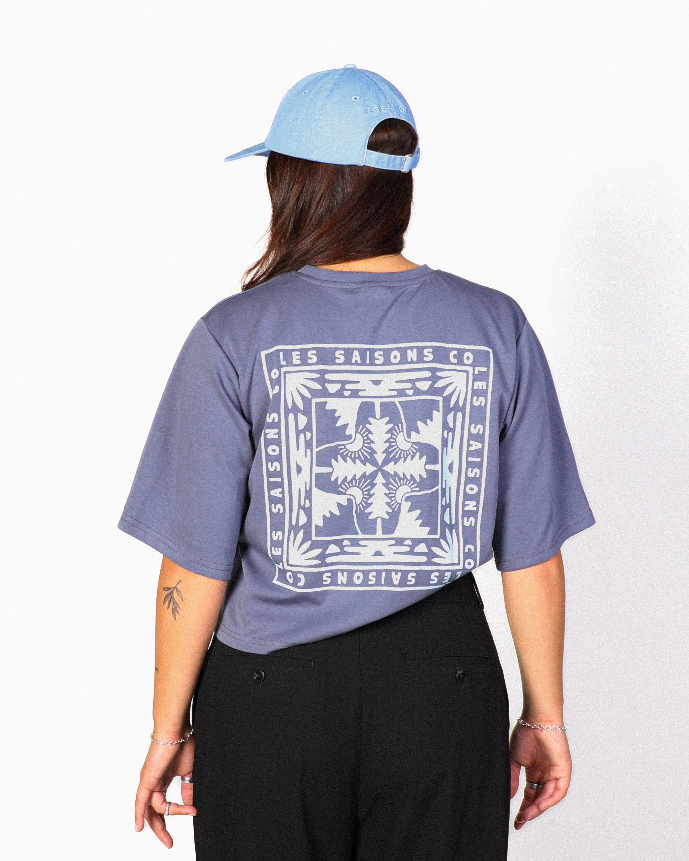 A person shown from the back wearing a light blue baseball cap, a slate blue graphic t-shirt with a large white bandana-style print, and black wide-leg pants, standing against a white studio background.