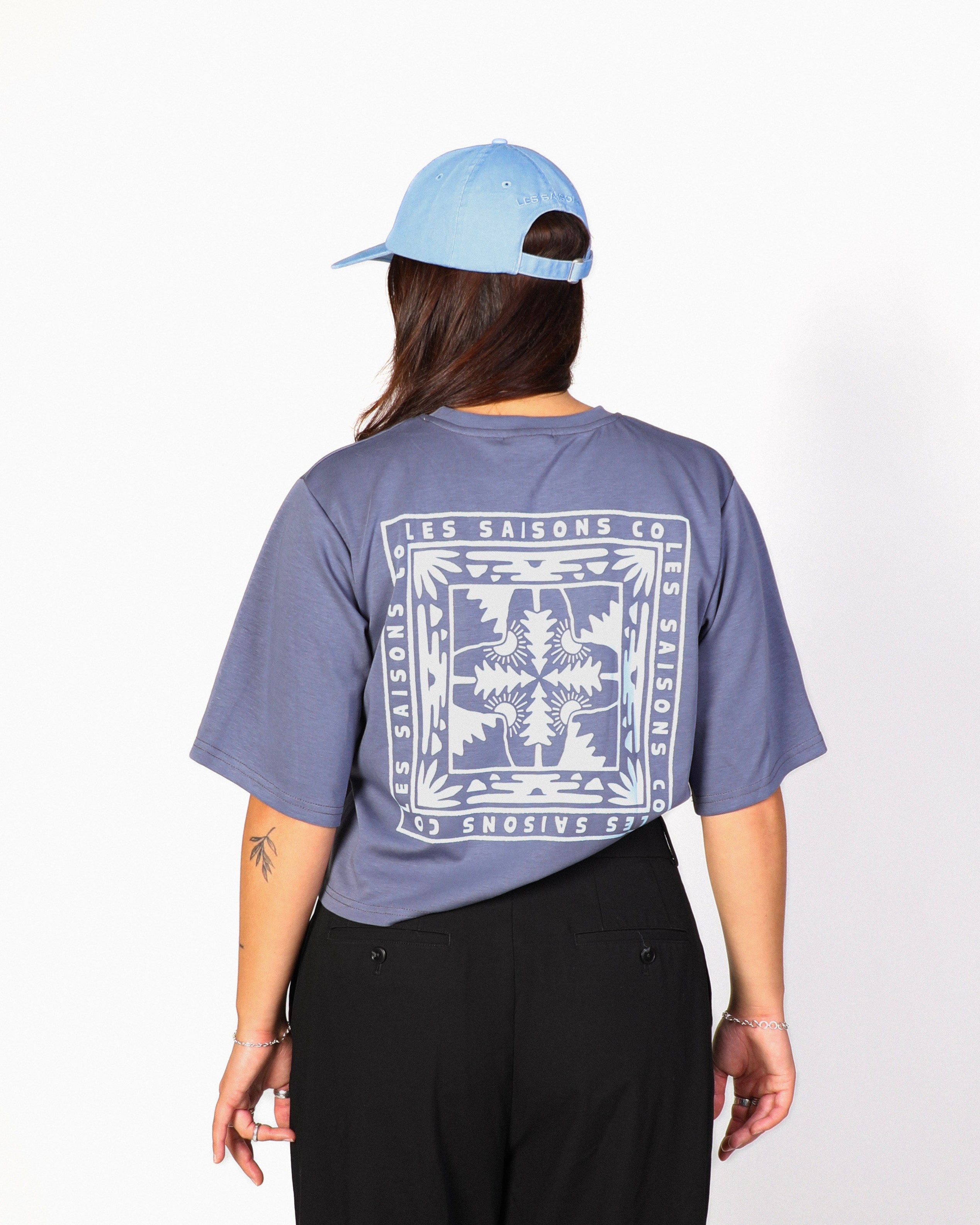 A person shown from the back wearing a light blue baseball cap, a slate blue graphic t-shirt with a large white bandana-style print, and black wide-leg pants, standing against a white studio background.