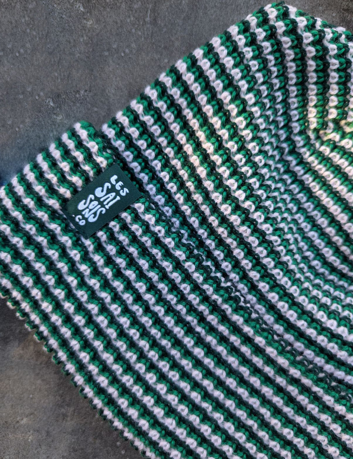 Zoom on the green beanie with a green les Saisons tag on a gray concrete surface, made in Canada.