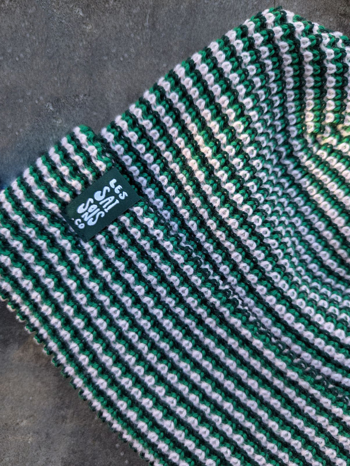 Zoom on the green beanie with a green les Saisons tag on a gray concrete surface, made in Canada.