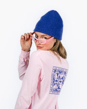 Person wearing a blue beanie and pink sunglasses with a pink sweatshirt featuring a logo on a white background
