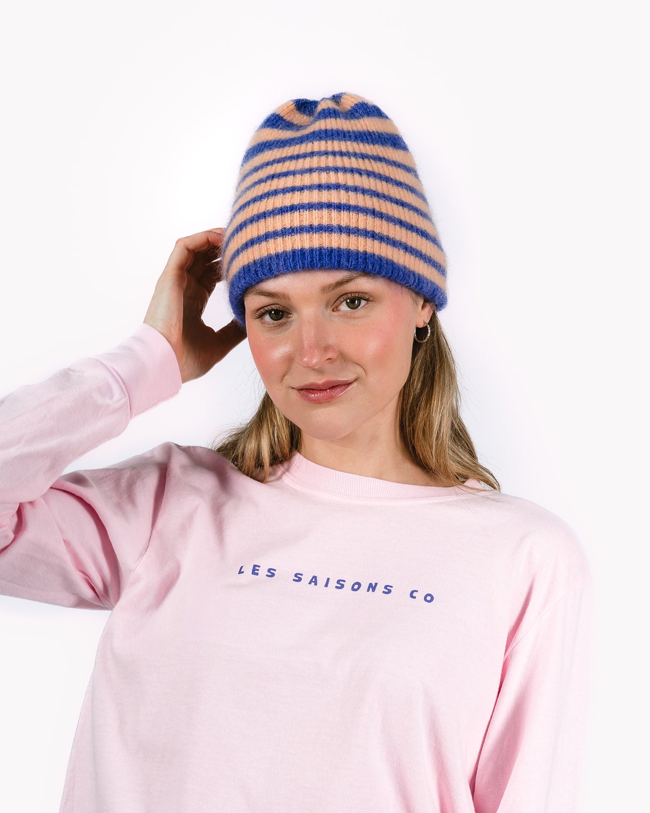 Person wearing a striped beanie and pink sweatshirt with 'LES SAISONS CO' text on a white background