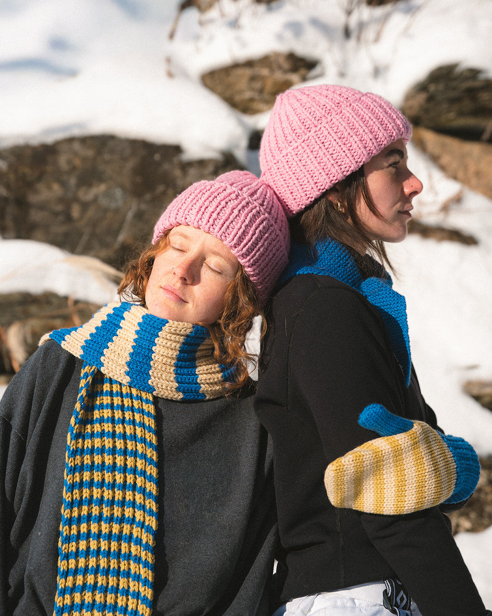 Two people wearing pink hats and colorful scarves standing in a snowy landscape.