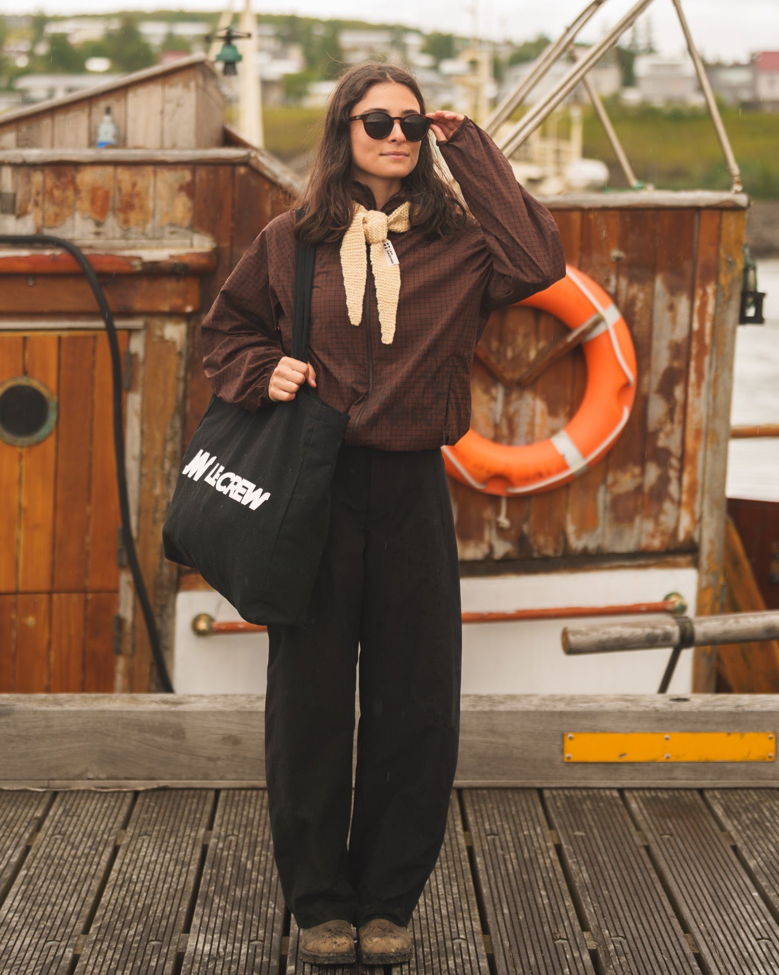 A woman stands on a wooden dock in front of a rustic boat, wearing a brown long-sleeve top with a cream neck bow, black wide-leg pants, and holding a black tote bag with white text.