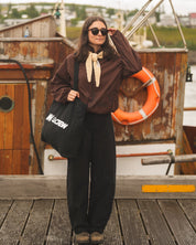A woman stands on a wooden dock in front of a rustic boat, wearing a brown long-sleeve top with a cream neck bow, black wide-leg pants, and holding a black tote bag with white text.