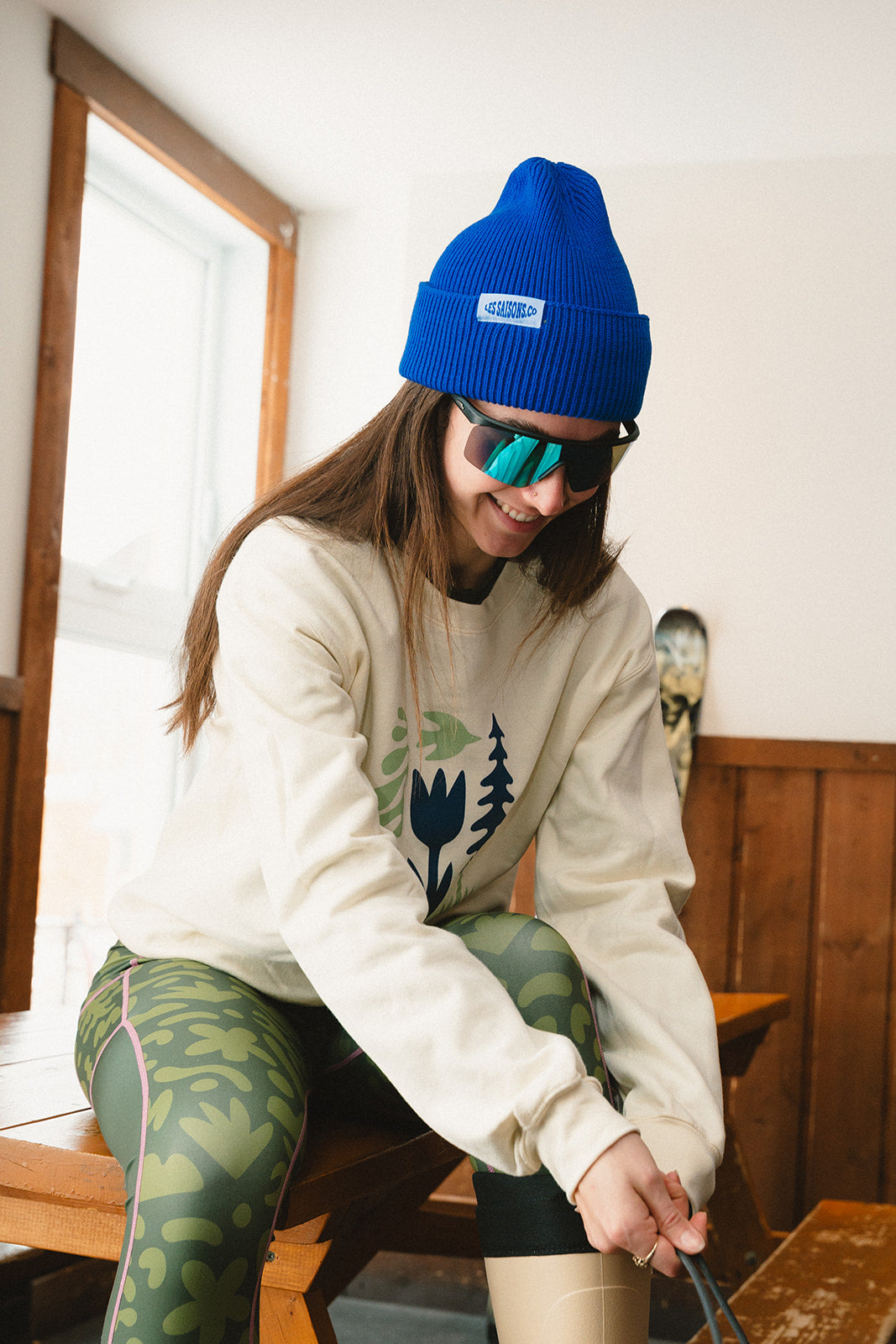 Person wearing a blue beanie and sunglasses, sitting indoors with skis in the background.