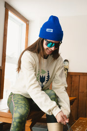 Person wearing a blue beanie and sunglasses, sitting indoors with skis in the background.