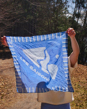 Person holding a blue and white patterned scarf bandana with 'Les Saisons' branding outdoors and a mountain