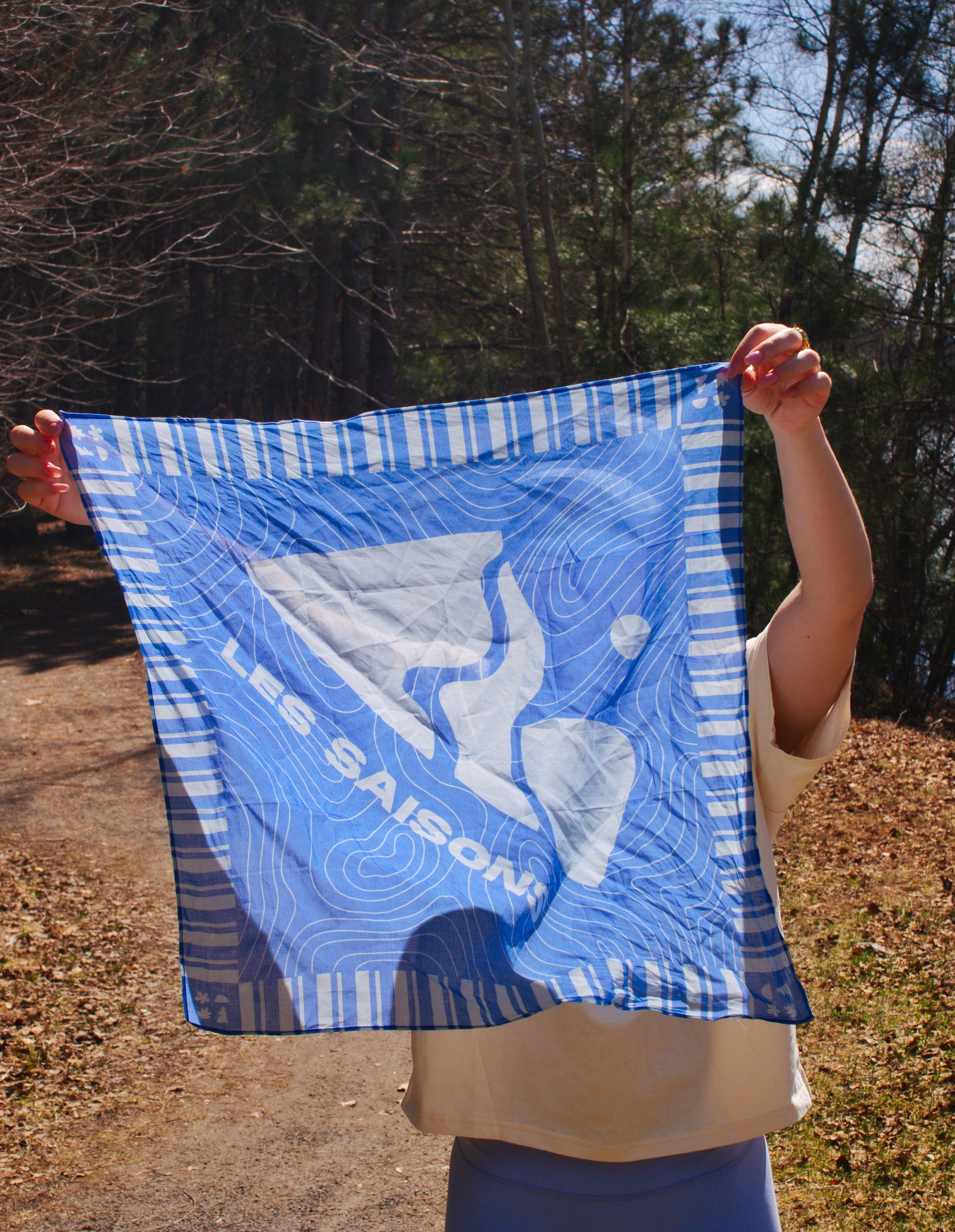 Person holding a blue and white patterned scarf bandana with 'Les Saisons' branding outdoors and a mountain