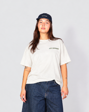 A model stands against a light gray studio background wearing a white graphic t-shirt, dark blue denim shorts, brown shoes, white socks, and a black baseball cap.