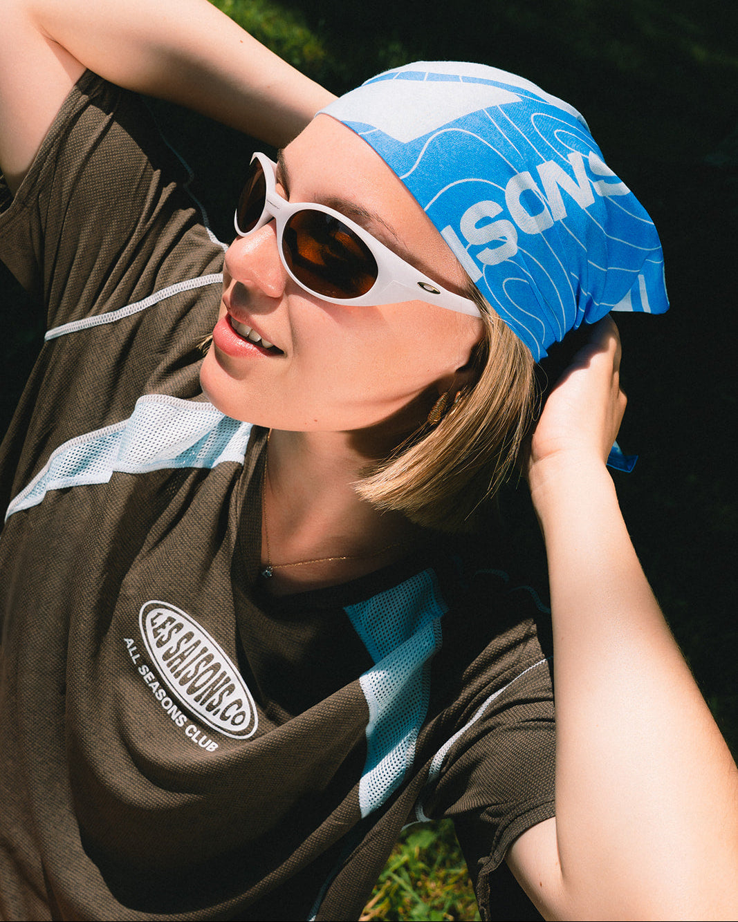 A woman outdoors wearing a brown active shirt, a blue and white patterned bandana, along with white-framed sunglasses.