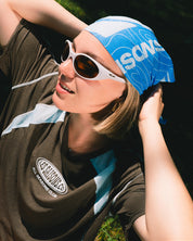 A woman outdoors wearing a brown active shirt, a blue and white patterned bandana, along with white-framed sunglasses.