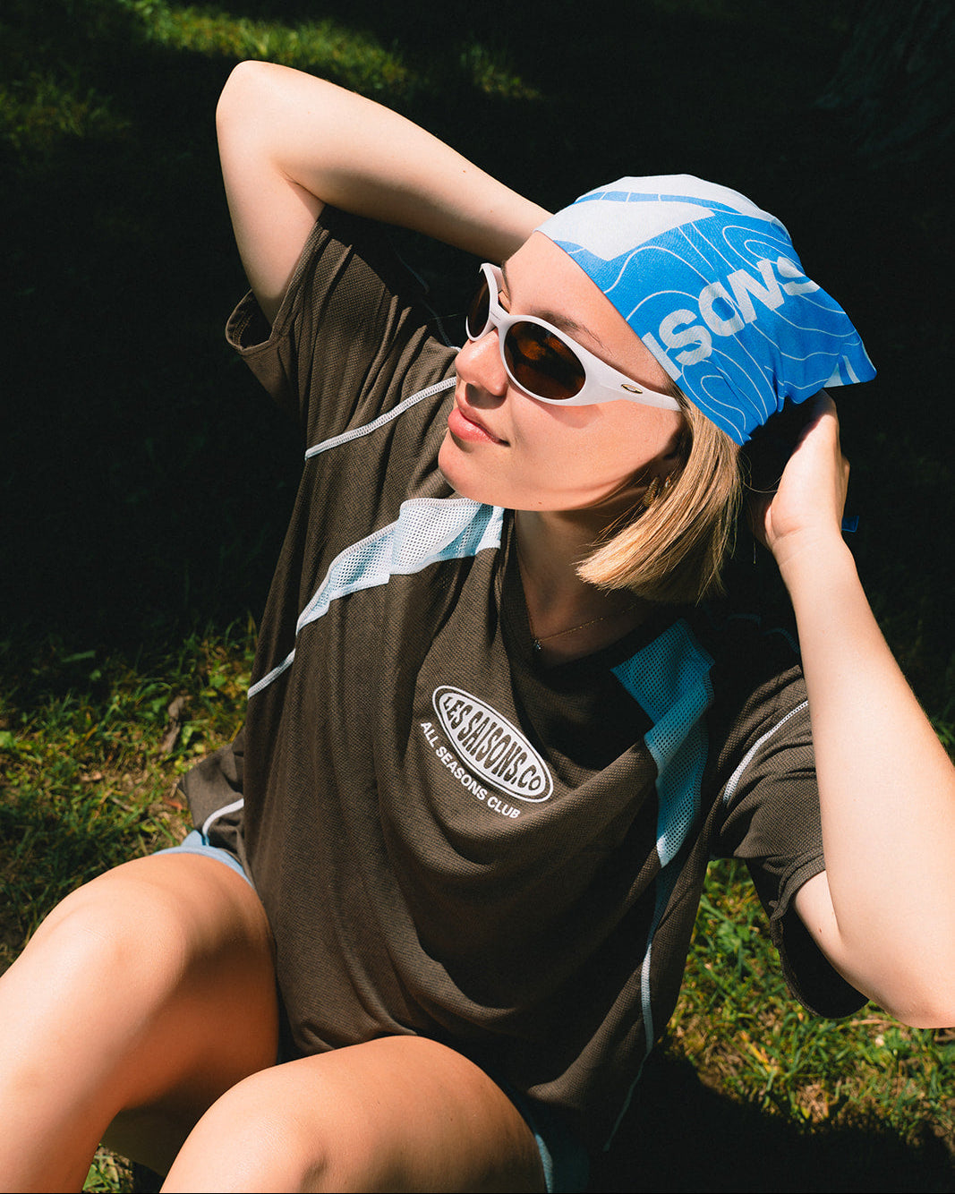 A woman sitting outdoors in the sun wearing a blue and white bandana with 'SONS' printed on it, white sunglasses, and a dark brown t-shirt with a circular logo.