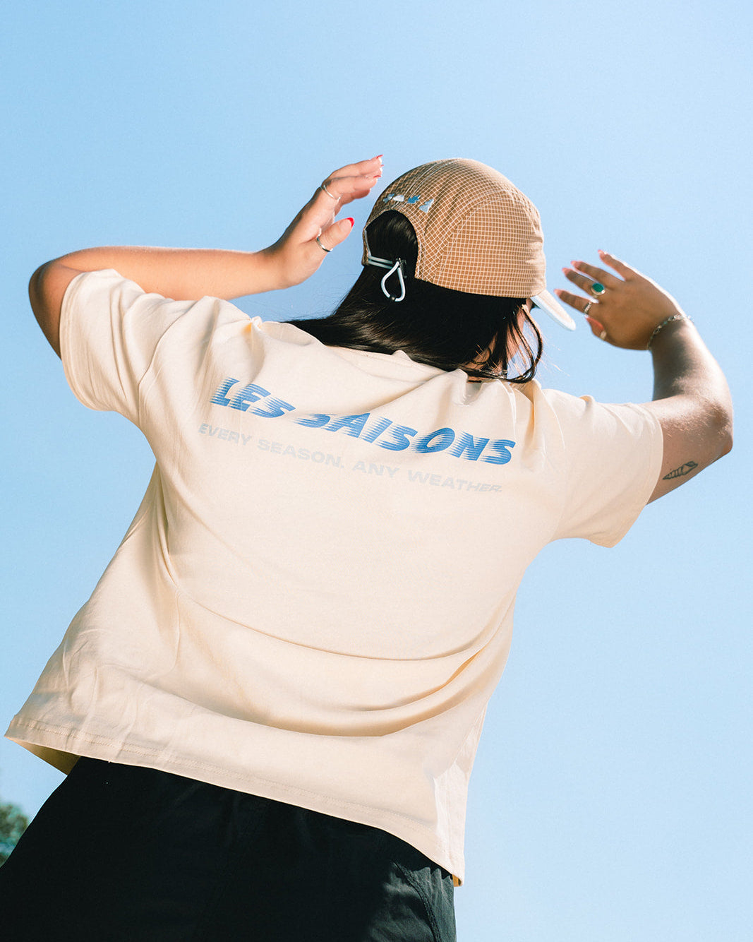 A person viewed from behind wearing a cream-colored t-shirt with blue 'LES SAISONS' text on the back, a beige baseball cap, and black pants, set against a clear blue sky.
