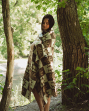 A woman wrapped in a brown and beige patterned blanket stands on a forest path between tree trunks, with green foliage overhead.