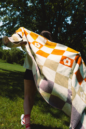 A person outdoors on grass under trees, wrapped in a large checkered blanket with orange, cream, beige, and muted purple squares and white flower motifs.