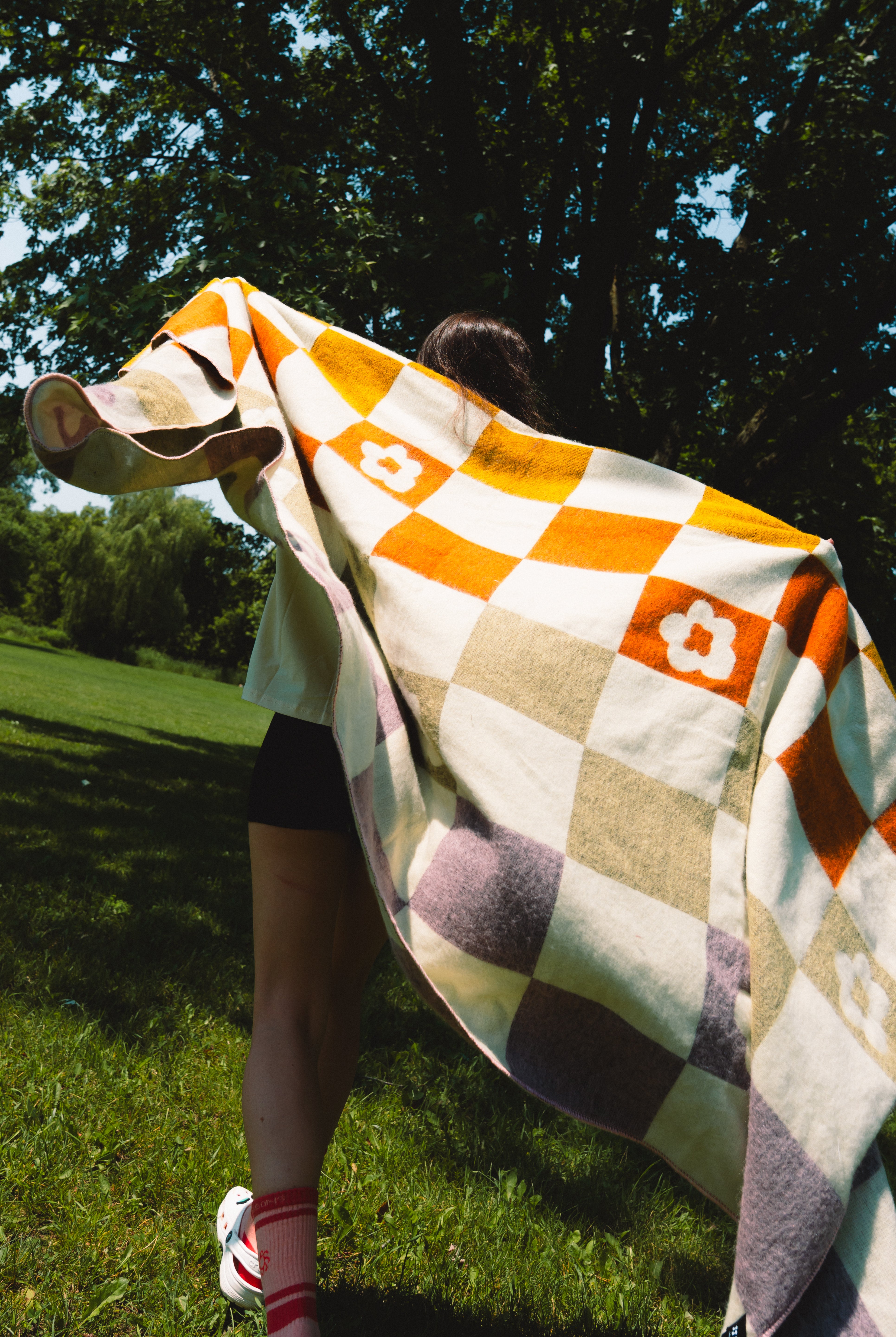 A person outdoors on grass under trees, wrapped in a large checkered blanket with orange, cream, beige, and muted purple squares and white flower motifs.