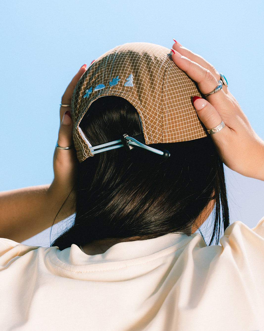 A person viewed from behind wearing a beige and brown grid-patterned cap, adjusting it with both hands. The person has long black hair and is wearing a cream-colored t-shirt with blue text that reads 'LES SAISONS'.
