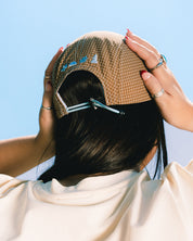 A person viewed from behind wearing a beige and brown grid-patterned cap, adjusting it with both hands. The person has long black hair and is wearing a cream-colored t-shirt with blue text that reads 'LES SAISONS'.