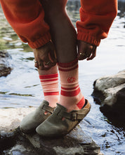 A person sitting on rocks by a body of water, wearing red and white striped crew socks with a small red sun motif, olive green clogs, and rust-orange sweatpants.