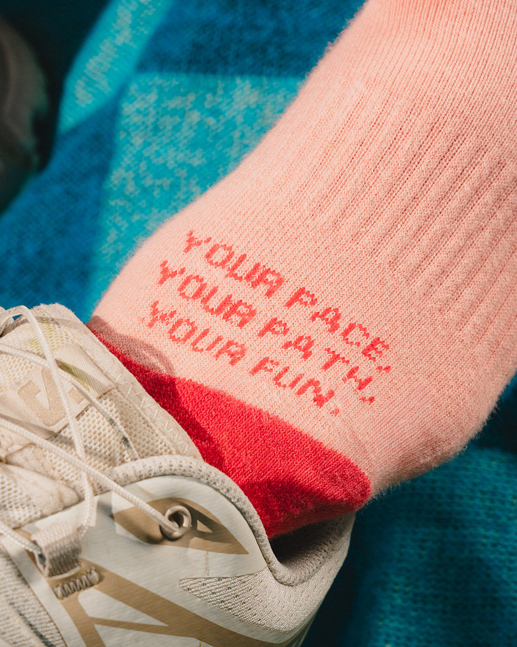 A close-up of a light pink athletic sock with red text reading 'YOUR PACE, YOUR PATH, YOUR FUN.' worn with a beige trail running shoe over a blue textured blanket.