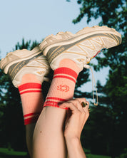 A person outdoors wearing chunky Salomon sneakers and bright pink crew socks with red stripes and a red logo, with their legs raised against a blurred park background.