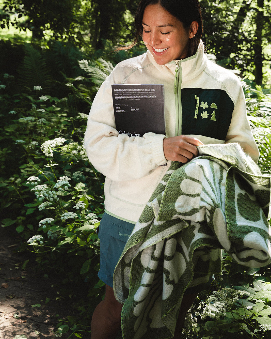 A woman standing outdoors in a wooded area, smiling while holding a cream zip-up fleece jacket with a green chest patch and a green patterned blanket.