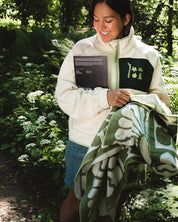 A woman standing outdoors in a wooded area, smiling while holding a cream zip-up fleece jacket with a green chest patch and a green patterned blanket.