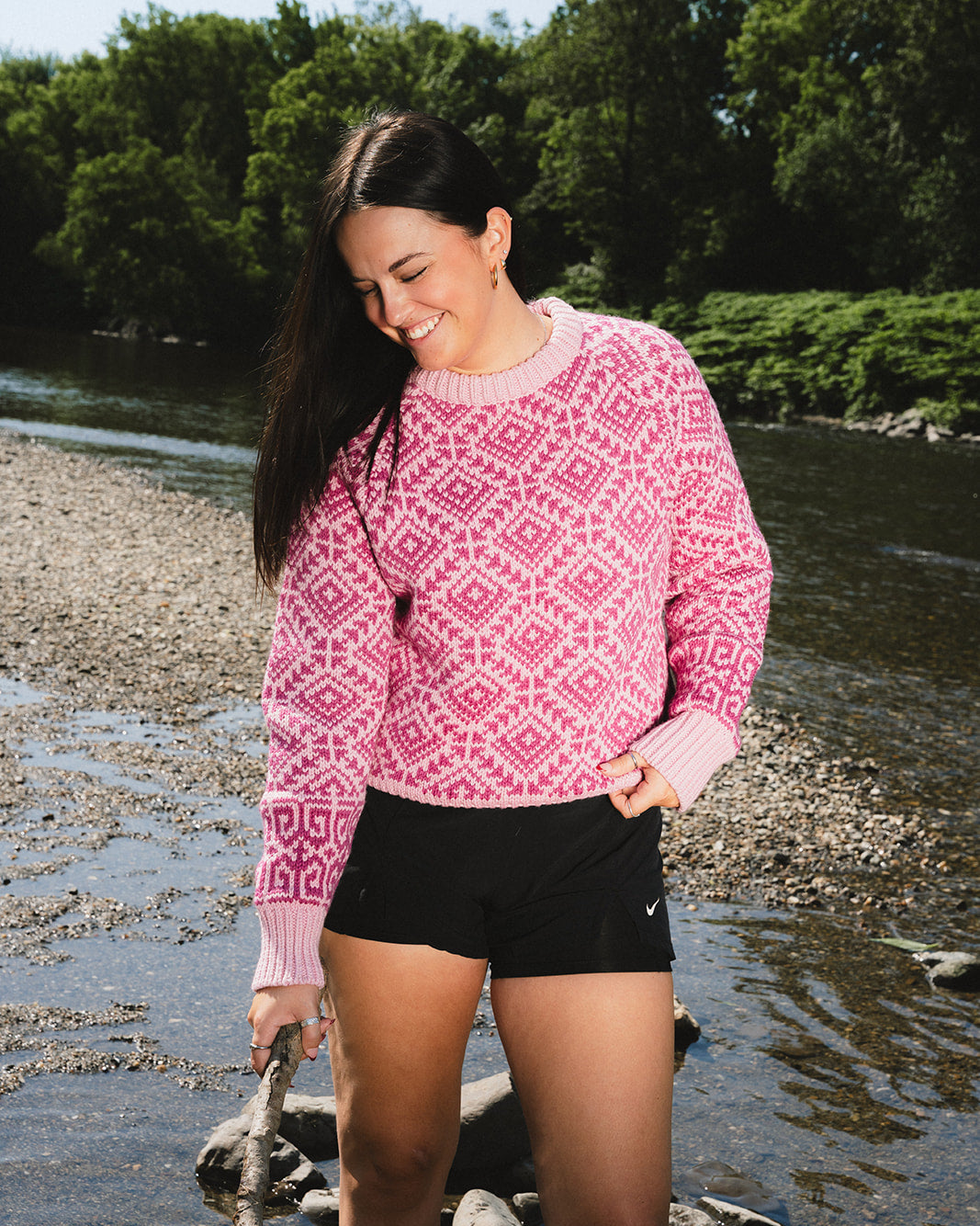 A woman standing in shallow water near a riverbank, wearing a pink and magenta geometric patterned knit sweater and black athletic shorts.