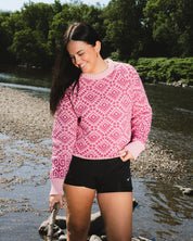 A woman standing in shallow water near a riverbank, wearing a pink and magenta geometric patterned knit sweater and black athletic shorts.