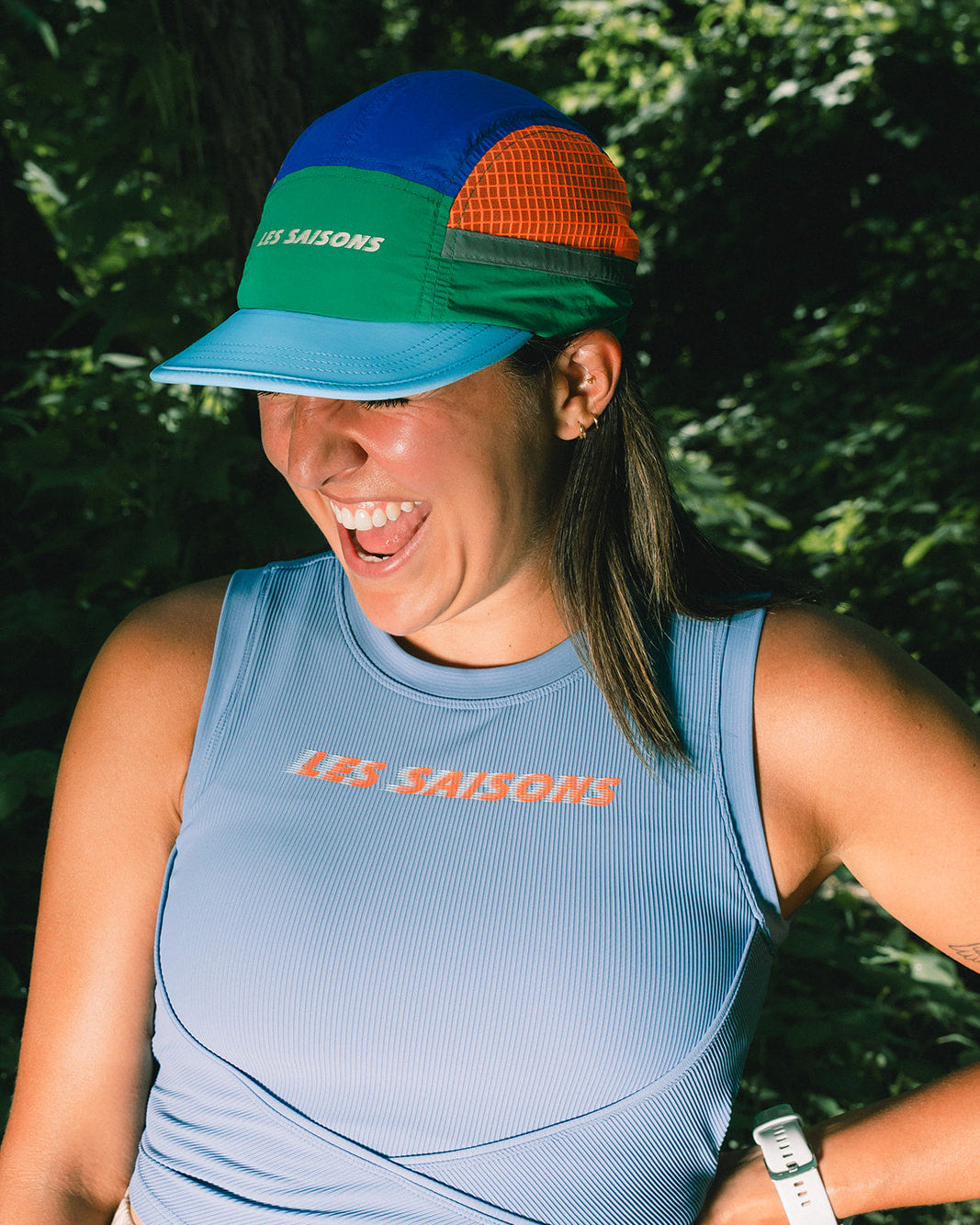 A woman outdoors wearing a light blue ribbed tank top and a multi-colored trucker cap with 'LES SAISONS' branding. The cap features blue, green, orange, and gray panels.