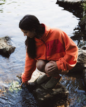 A woman crouches in shallow water near rocks, wearing a bright orange and red fleece pullover, brown boots, and dark leggings.