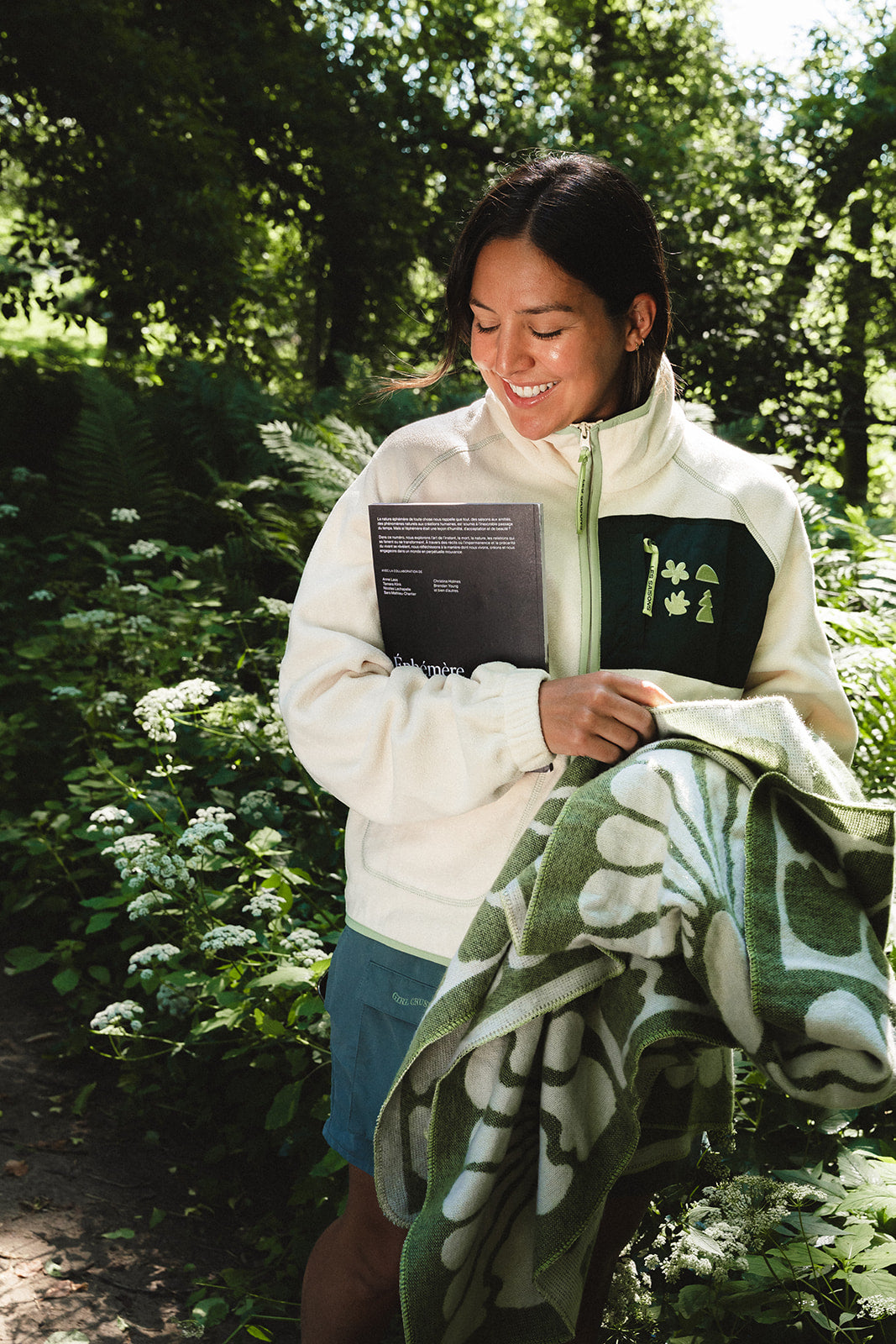 A woman standing outdoors in a wooded area, smiling while holding a cream zip-up fleece jacket with a green chest patch and a green patterned blanket.