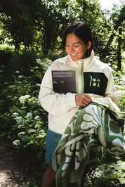A woman standing outdoors in a wooded area, smiling while holding a cream zip-up fleece jacket with a green chest patch and a green patterned blanket.