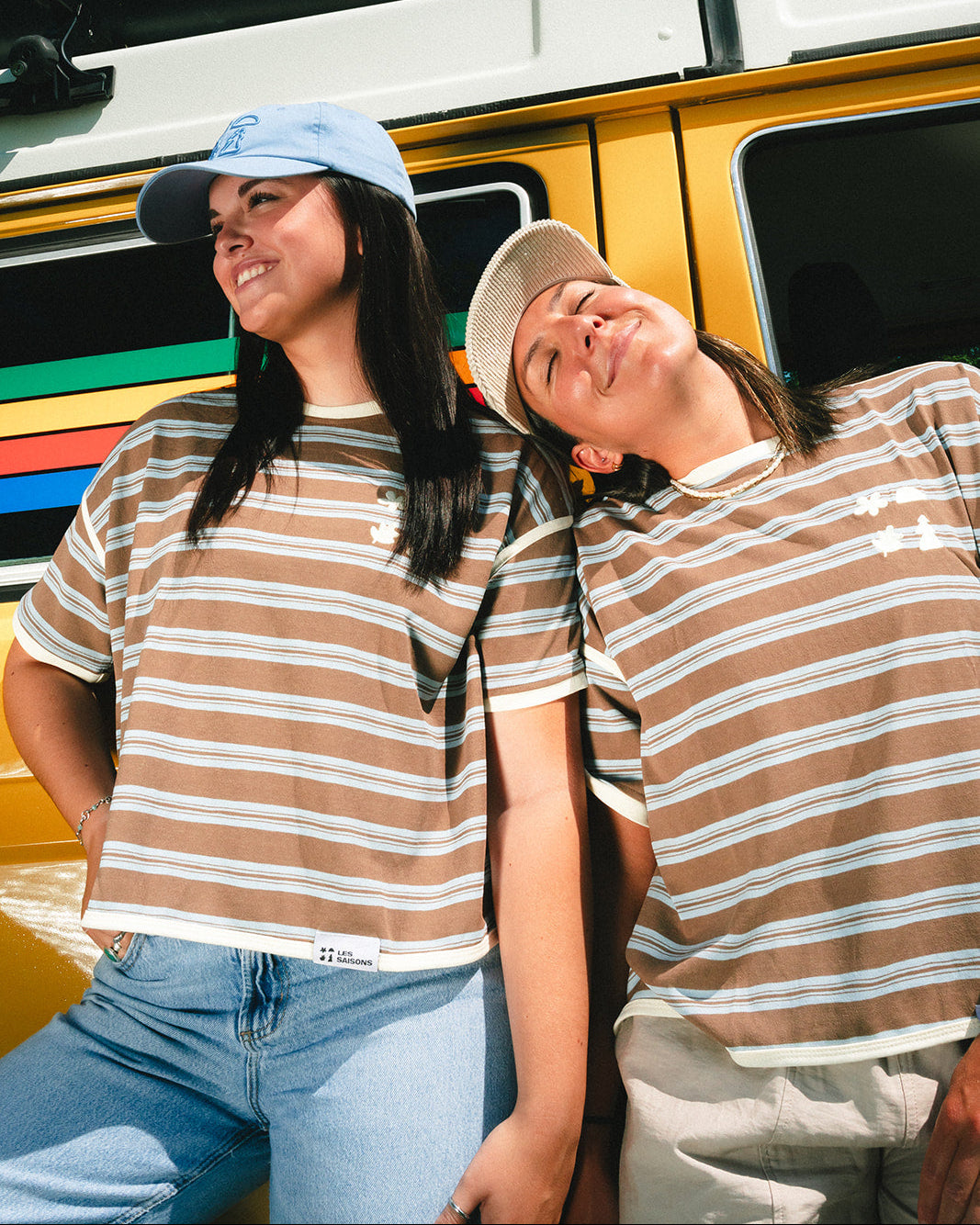 Two young women wearing matching brown and white striped t-shirts and baseball caps, posing outdoors in front of a yellow vehicle.