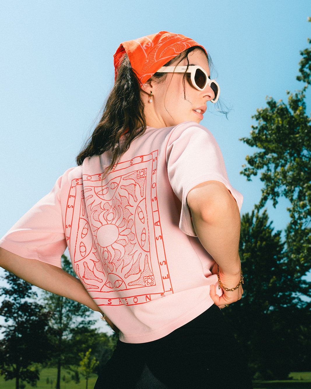 A woman outdoors wearing a light pink t-shirt with a large red mandala graphic on the back, paired with a bright orange bandana, white sunglasses, and a black skirt.