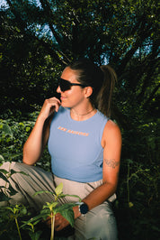 A woman wearing a light blue sleeveless crop top with 'LES SAISONS' printed in orange across the chest, paired with beige pants, sitting outdoors in a lush green environment.