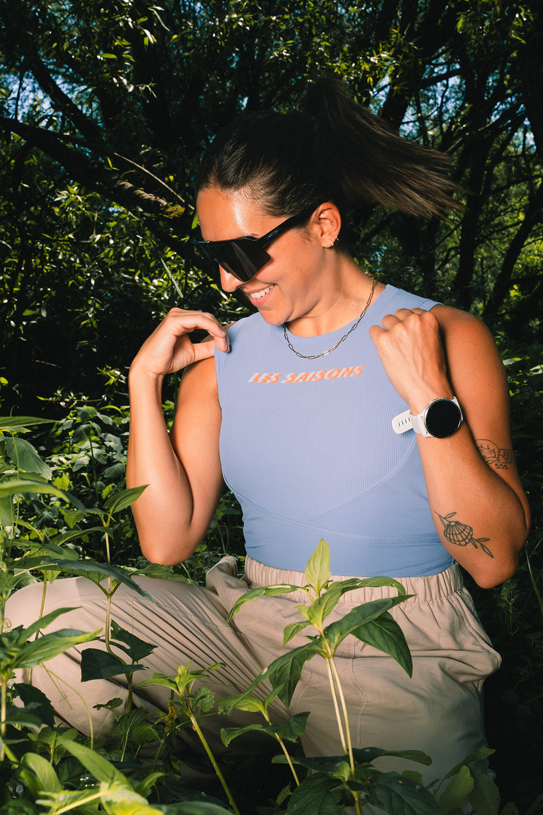 A woman wearing a light blue sleeveless top with 'LES SAISONS' printed in orange on the chest, paired with beige pants, posing outdoors among green plants and trees.