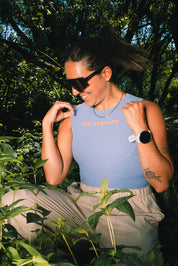 A woman wearing a light blue sleeveless top with 'LES SAISONS' printed in orange on the chest, paired with beige pants, posing outdoors among green plants and trees.