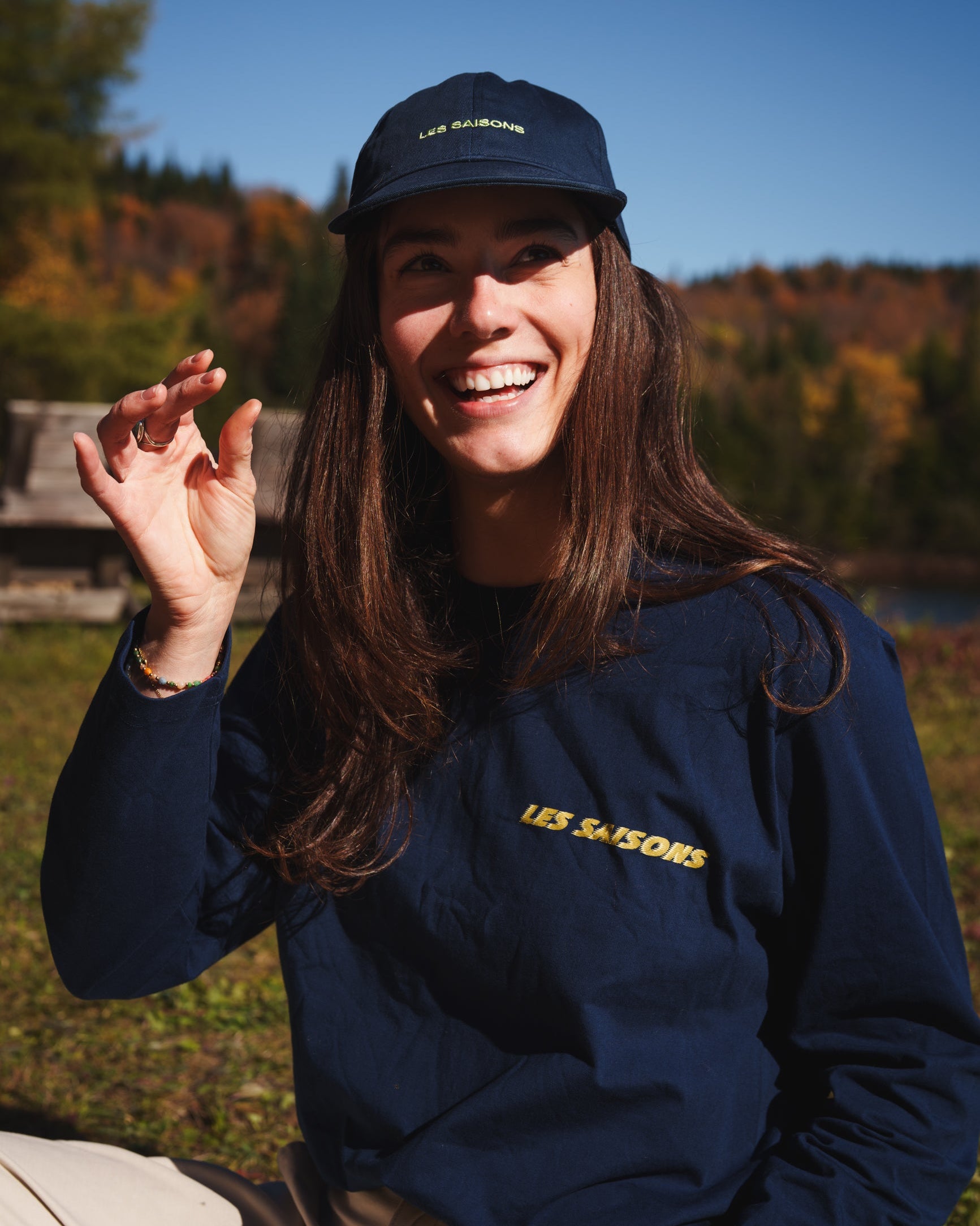 Person smiling outdoors in the navy Les Saisons Classic Longsleeve with yellow chest lettering and a matching navy cap
