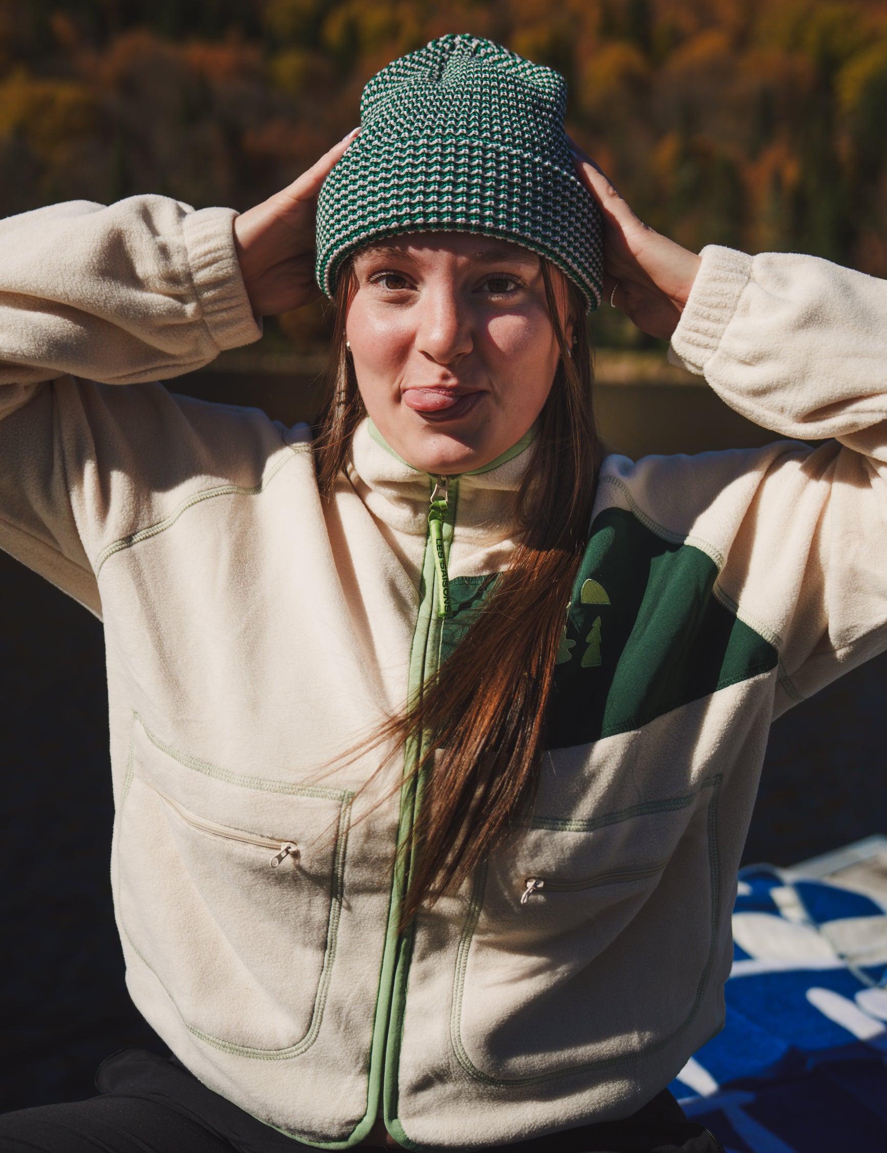 Person wearing the LesSaisons Green Lines beanie in green and white knit, holding both hands to the hat outdoors.