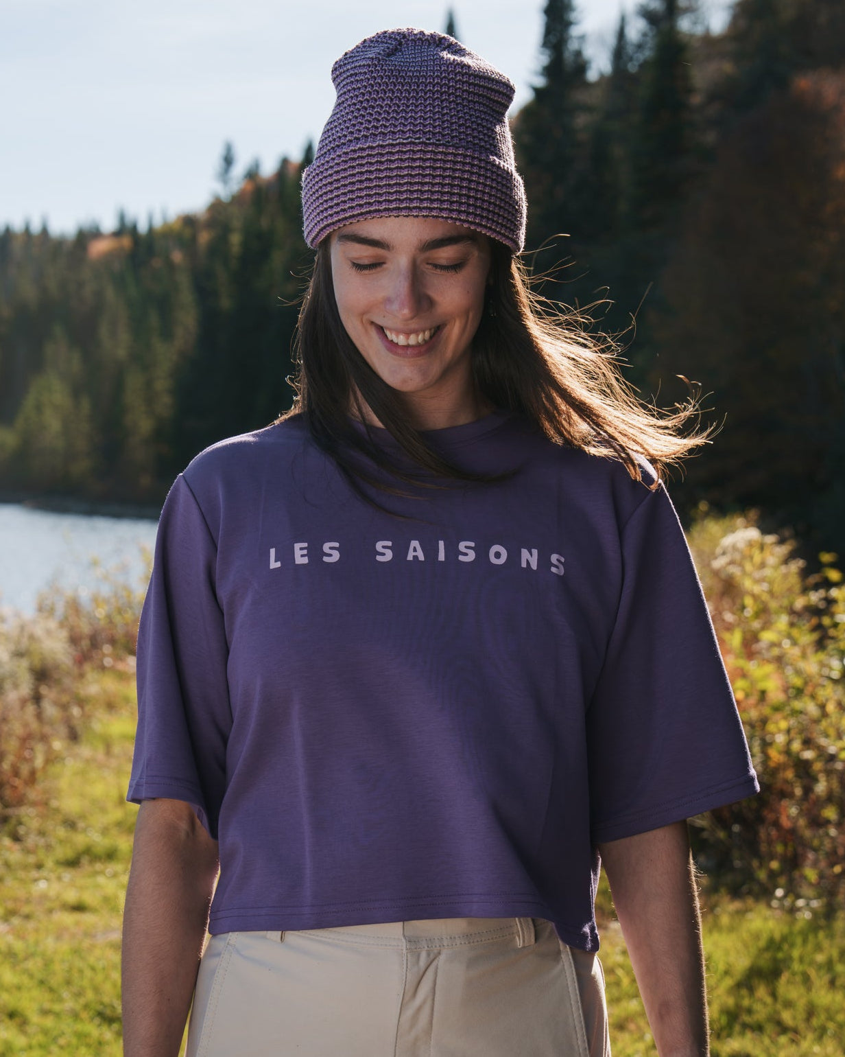 Person wearing the Les Saisons Purple Lines knit beanie with a folded cuff, smiling and looking down beside a lake