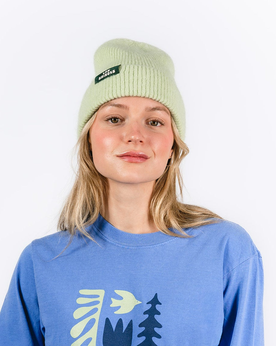 Person wearing a blue sweatshirt with 'Les Saisons' design and a light green Mohair beanie on a white background