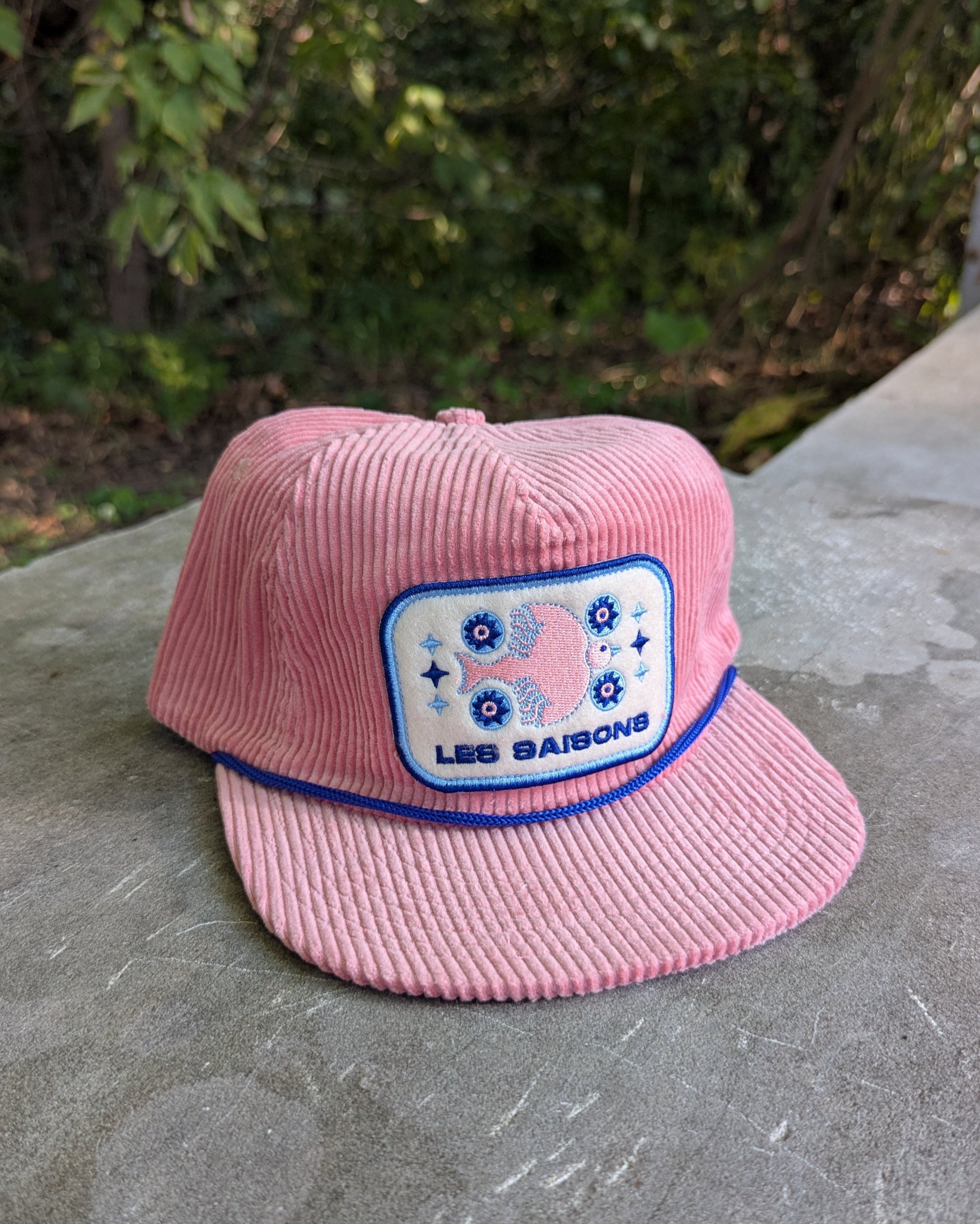 Pink cap with Artwork from Marie-Piel Artist on a concrete surface with greenery in the background