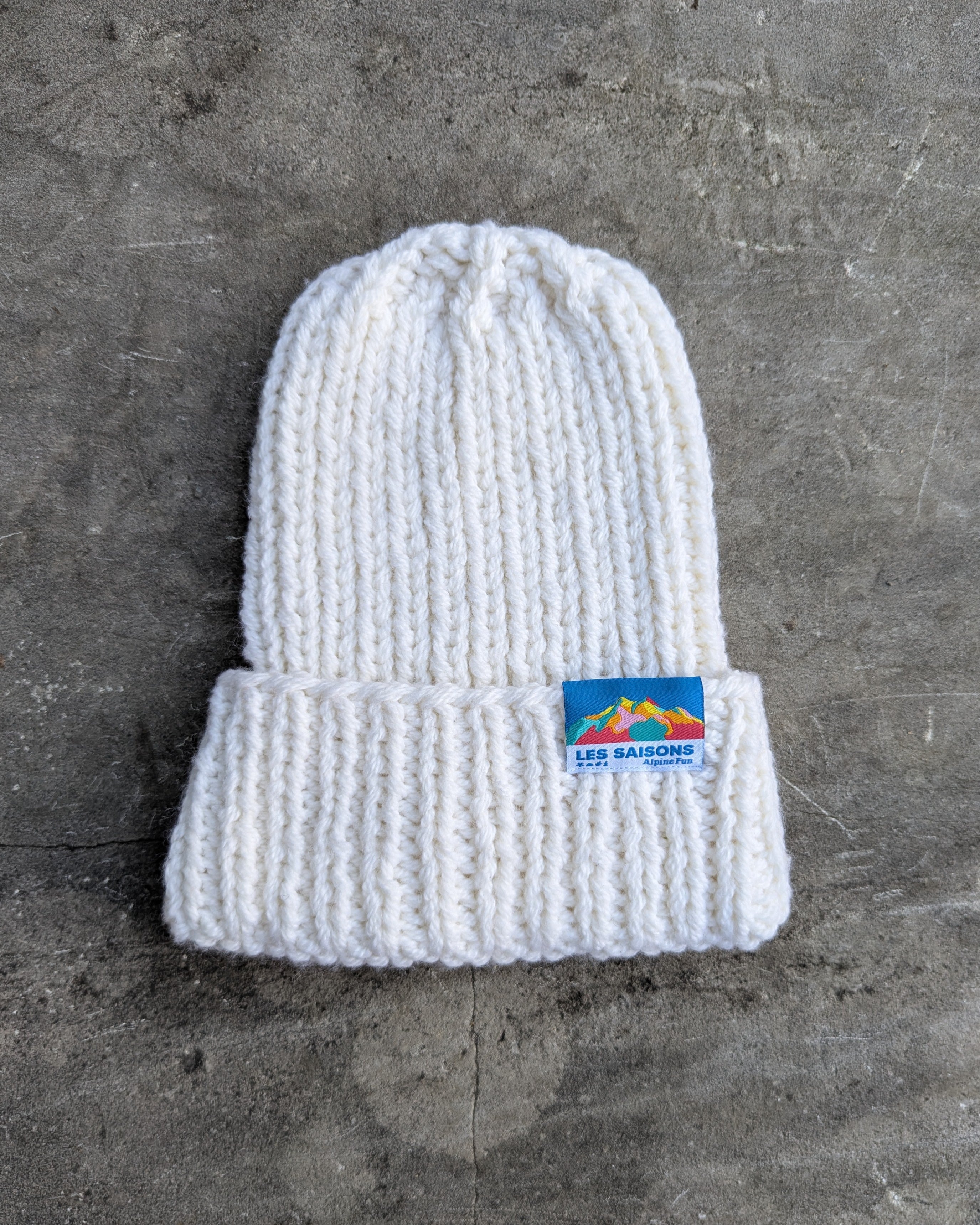 White knit beanie with a les Saisons logo on a gray surface