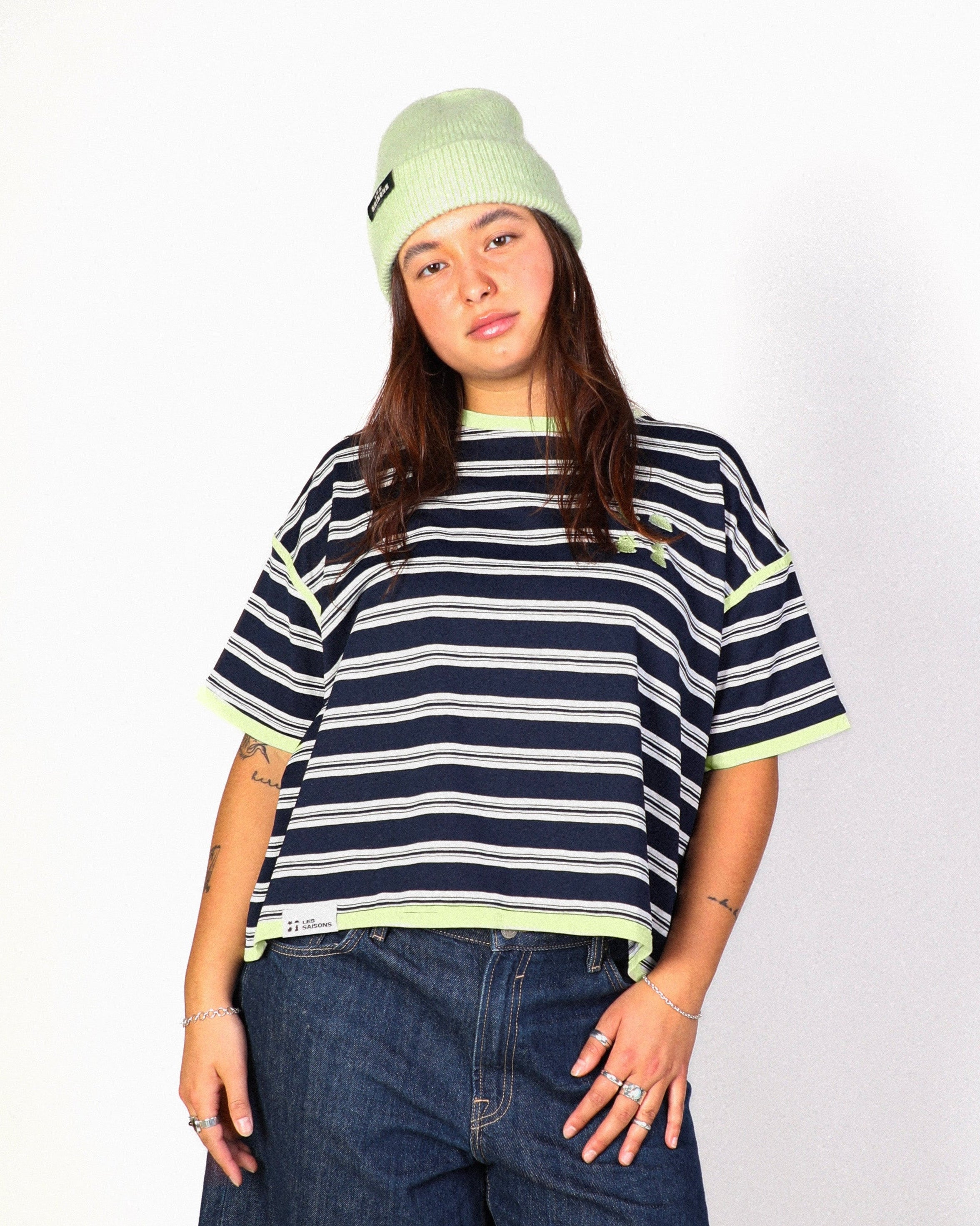 A model stands against a white studio background wearing a navy and white striped t-shirt with neon yellow-green trim, dark blue denim shorts, a light green beanie, white socks, and brown suede shoes.
