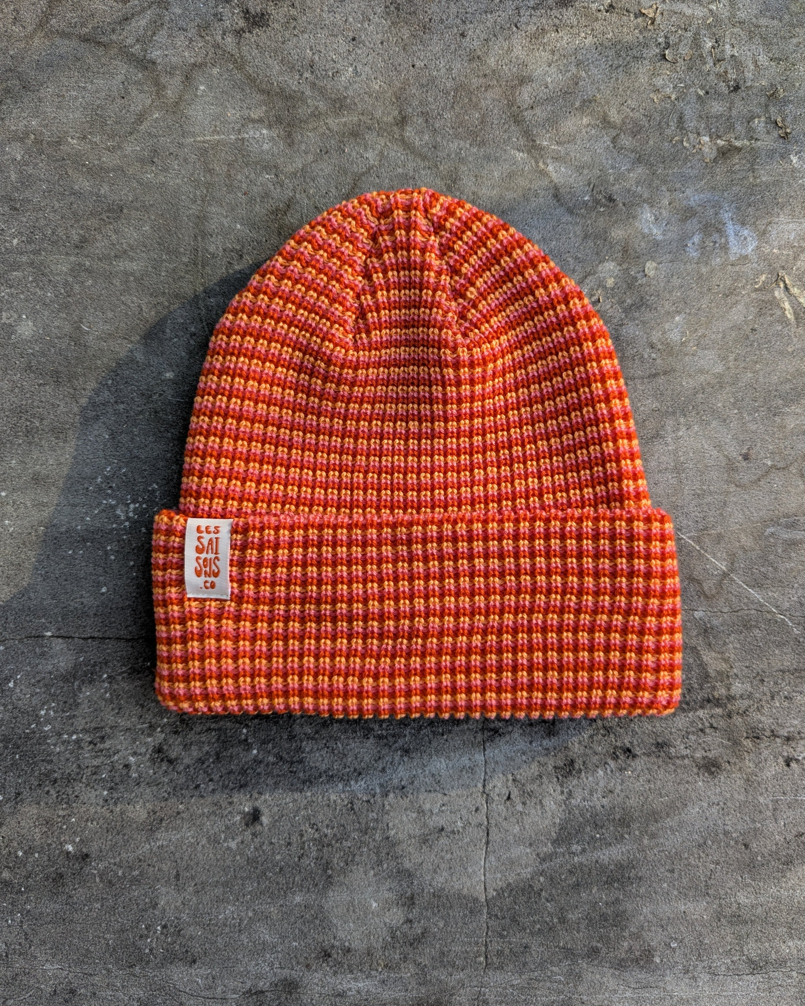 Orange beanie with a les Saisons tag on a gray concrete surface, made in Canada.