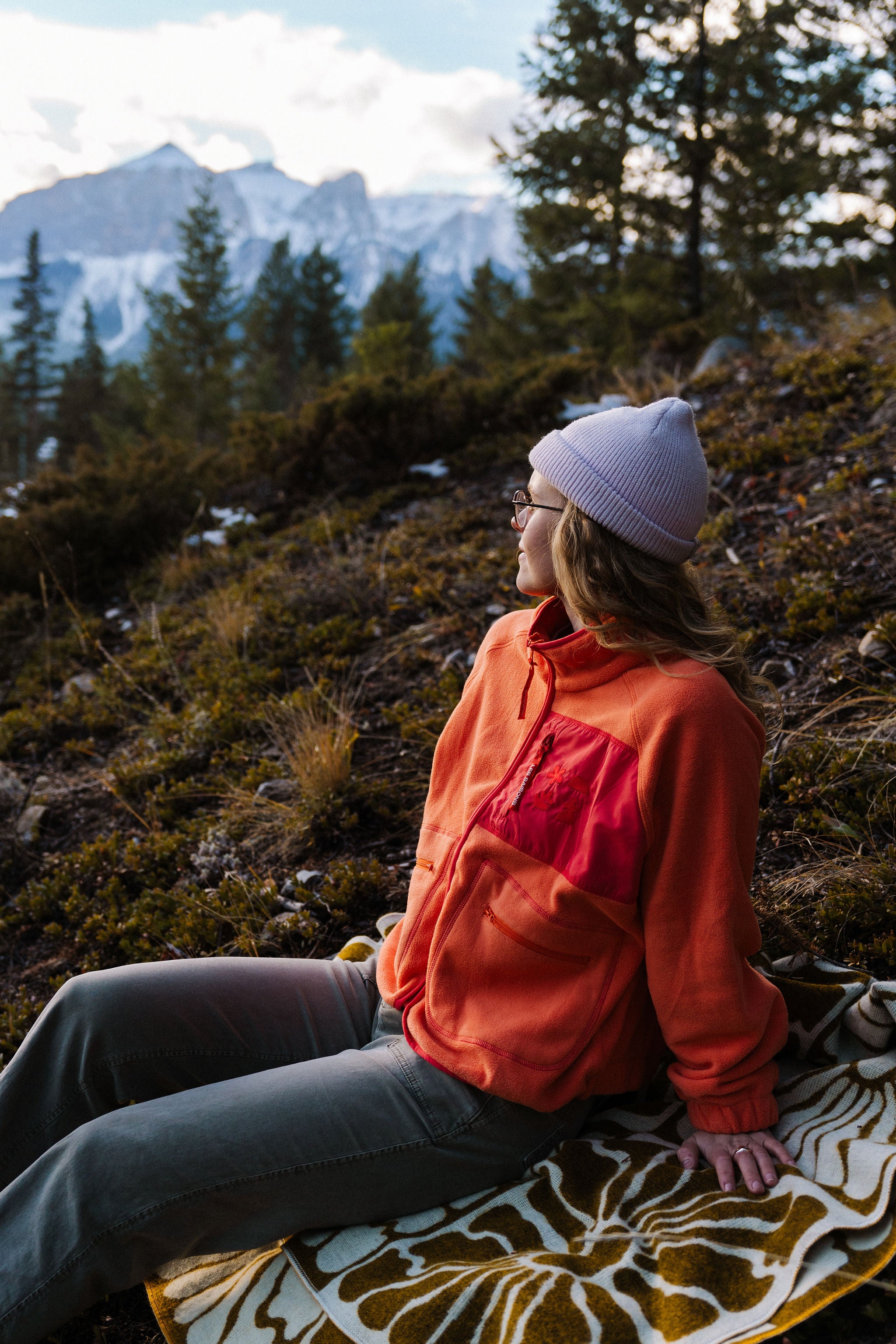 Person sitting on a blanket in a mountainous area wearing an orange fleece jacket les Saisons
