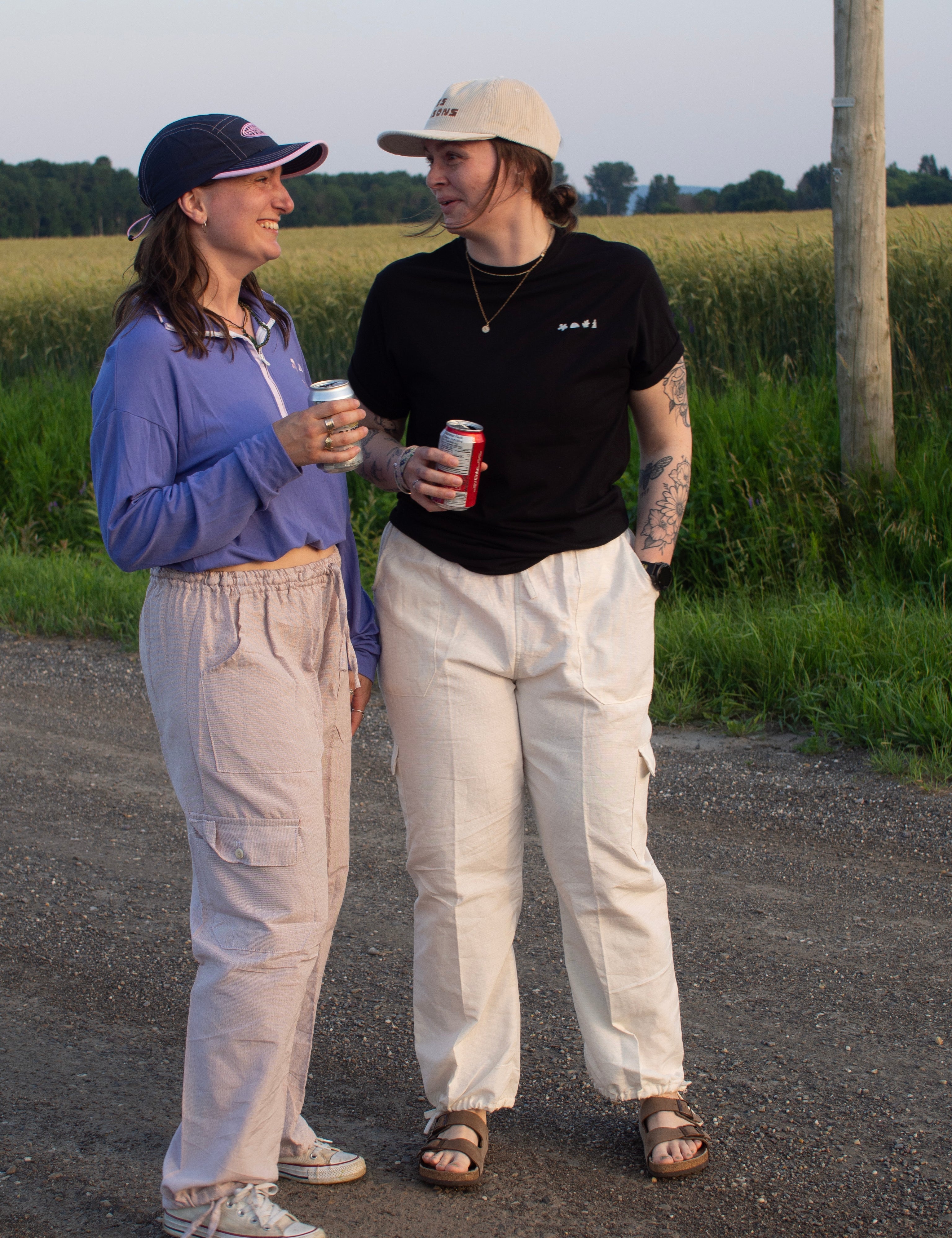 Two people standing on a road near a field, holding drinks and smiling.